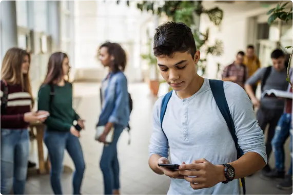 Jeune regardant un téléphone intelligent dans un couloir d’école avec d’autres adolescents en arrière-plan.