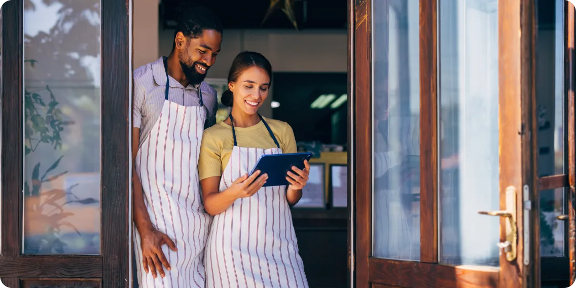 Deux propriétaires de petites entreprises, portant un tablier et souriants, regardent une tablette devant leur boutique.