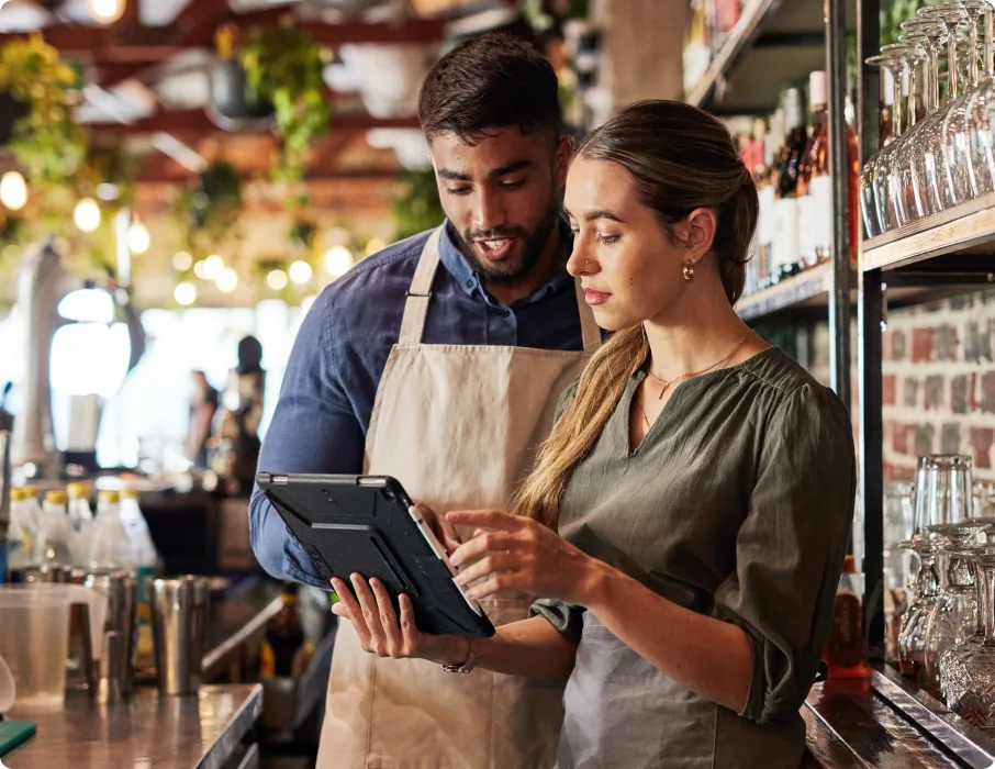 Two restaurant workers in aprons collaborating while using tablet device in rustic café interior