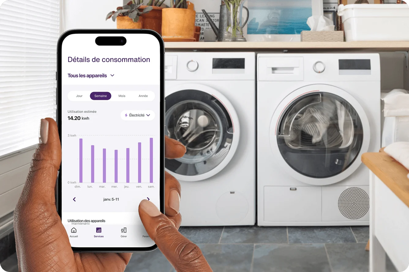 Une main tient un téléphone intelligent montrant la consommation d’énergie de l’appareil près de la laveuse et de la sécheuse