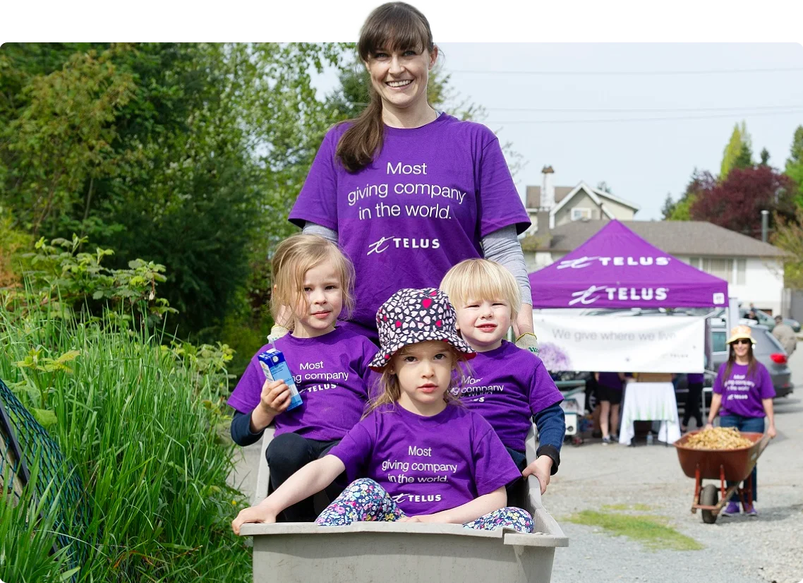 A TELUS volunteer standing behind three children seated in a wheelbarrow.