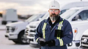 Man wearing blue coveralls and white hard hat holding a tablet, Standing in front of white fleet vehicles.