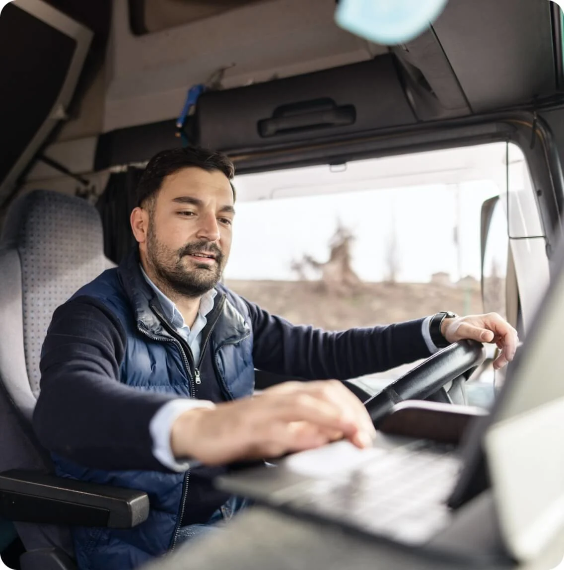A man in the driver's seat of a truck, working on a laptop, focused on his task.