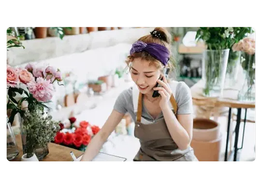 A smiling florist in a gray apron talks on the phone and writes on a clipboard. The shop is filled with potted plants and flowers. 