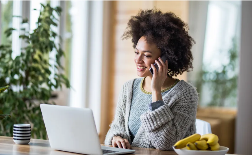 Woman using mobile phone in front of open laptop. Her coffee cup, plants and a bowl of bananas are visible in the frame. 