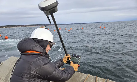 Worker in a hard hat using surveying equipment by the ocean with orange buoys in the water