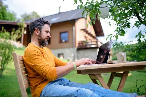 A man using his laptop while sitting on chair outside his home.
