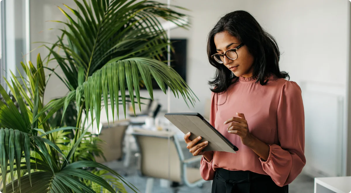 Businesswoman in a pink blouse, standing near a large potted plant, using a digital tablet in a modern office environment.