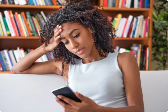 A seated woman viewing a smartphone with a bookshelf in the background.