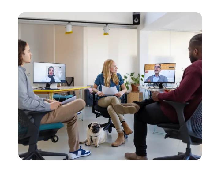 Three people and a dog (pug) seated in an office with two additional people on video screens.