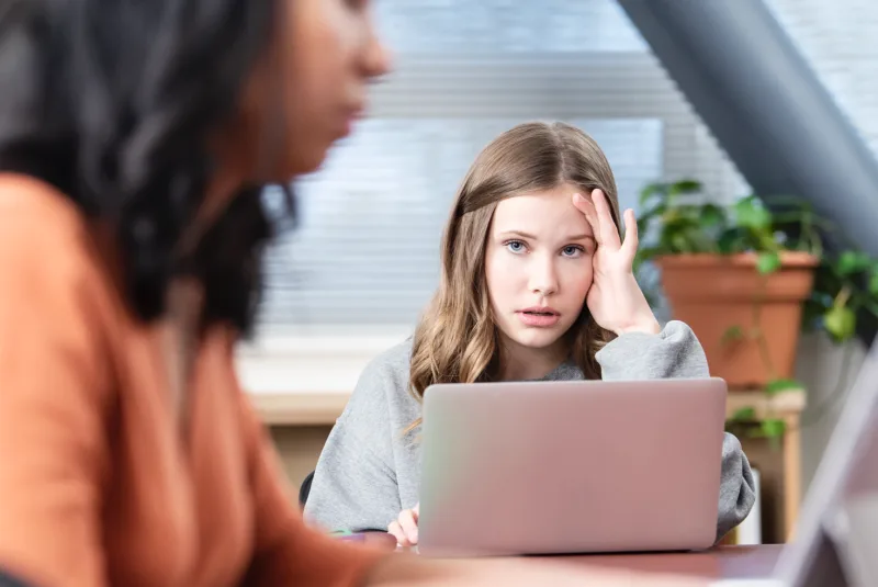 A teenage girl glances up from her laptop at another woman on her laptop.