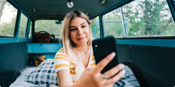 A woman sitting inside a camper van in a wooded area, with her arm outstretched and holding a smartphone to make a video call.