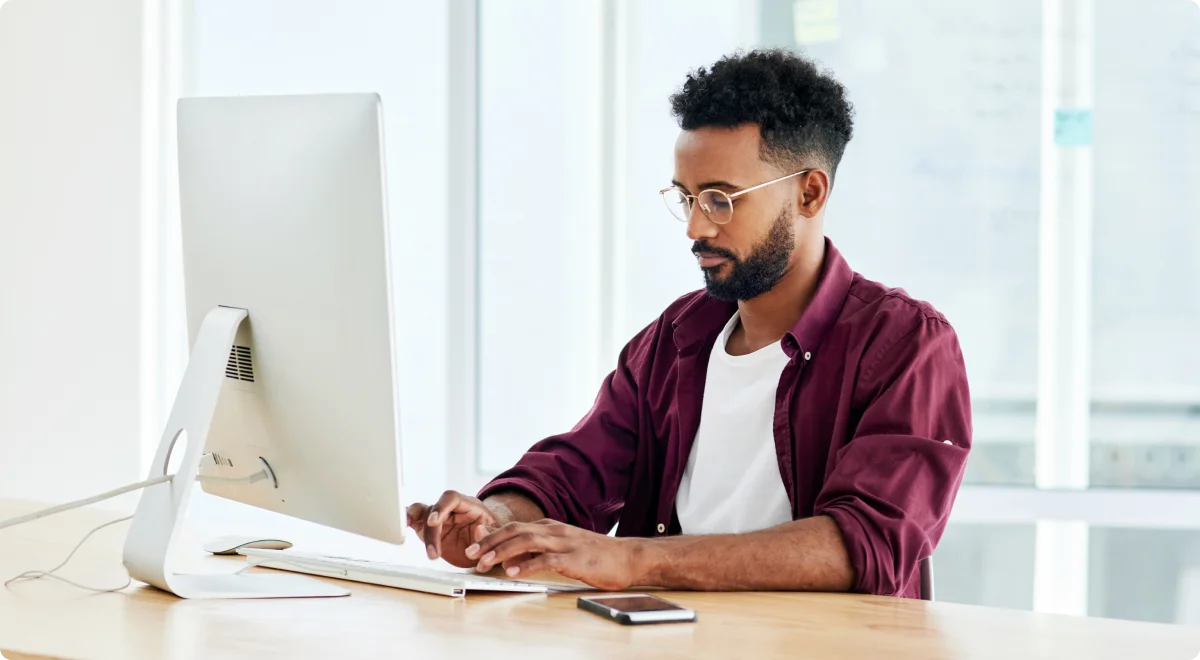 Young professional in a maroon shirt working on a desktop computer at a modern office desk, with a smartphone placed nearby.