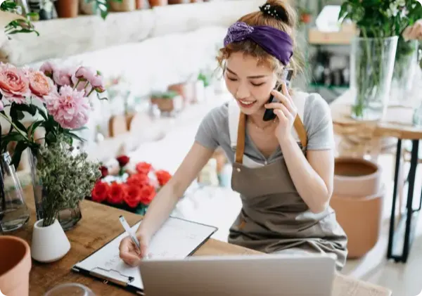 A smiling florist in a gray apron talks on the phone and writes on a clipboard. The shop is filled with potted plants and flowers.