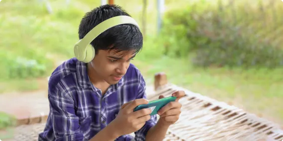 Un jeune garçon est assis dans un paysage verdoyant, porte des écouteurs jaunes et utilise une console de jeu vidéo.