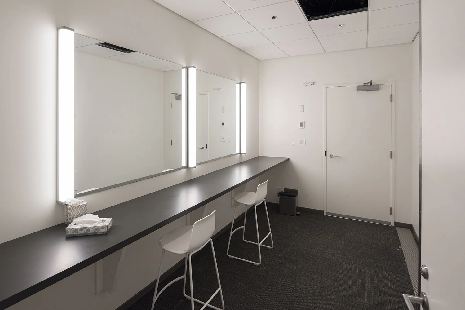 Green room with lit up mirrors, vanity and white chairs