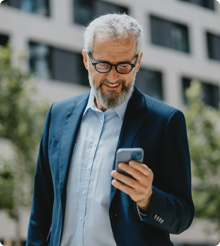 Mature businessman in glasses looks down at his phone while holding a coffee outside.