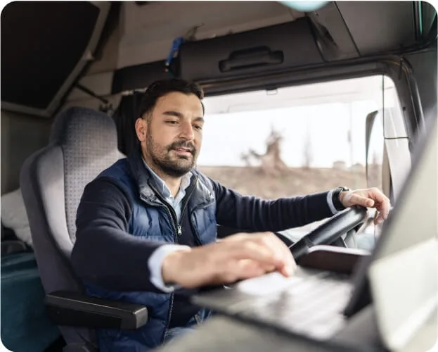 A man in the driver's seat of a truck, working on a laptop, focused on his task.