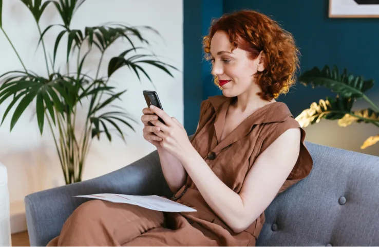 A woman sitting on a sofa is looking at a mobile device and smiling.