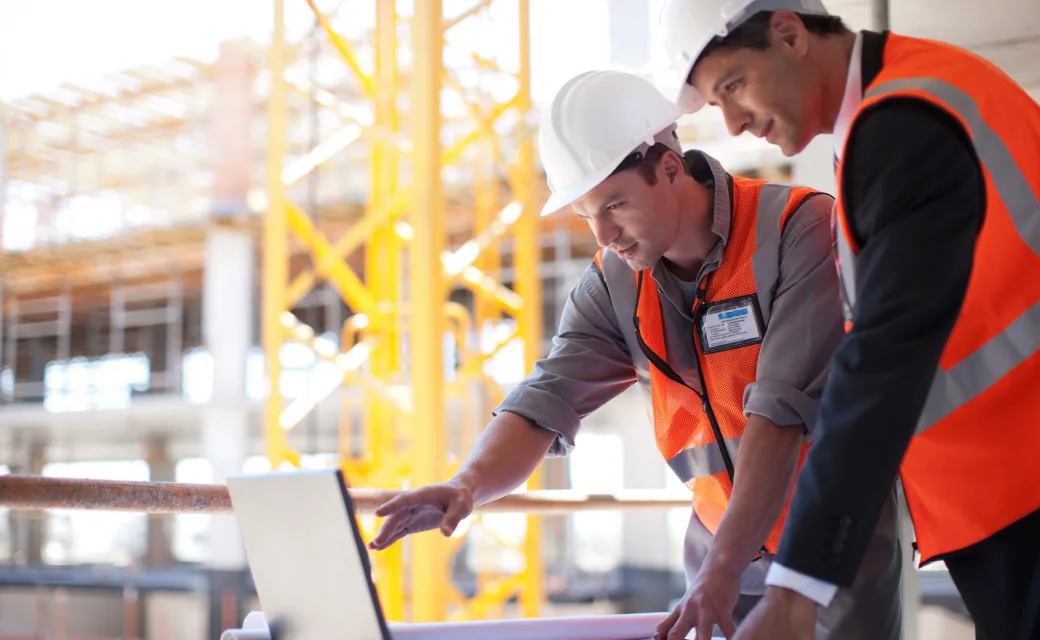 Two connected workers in white hard hats and orange safety vests discussing work over a laptop