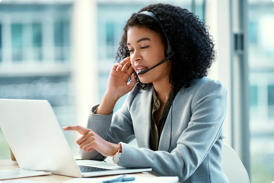 Business professional wearing a headset, working on a laptop in a modern office setting, engaging in a call while pointing at the screen.