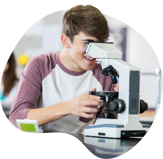 A teenager in a classroom examining an object through a microscope.
