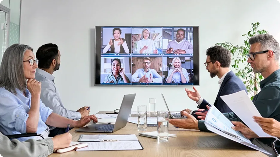 A building development team sits around a large table having an online meeting with a group of stakeholders.