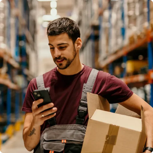 A warehouse worker carries a box and checks his smartphone between aisles.