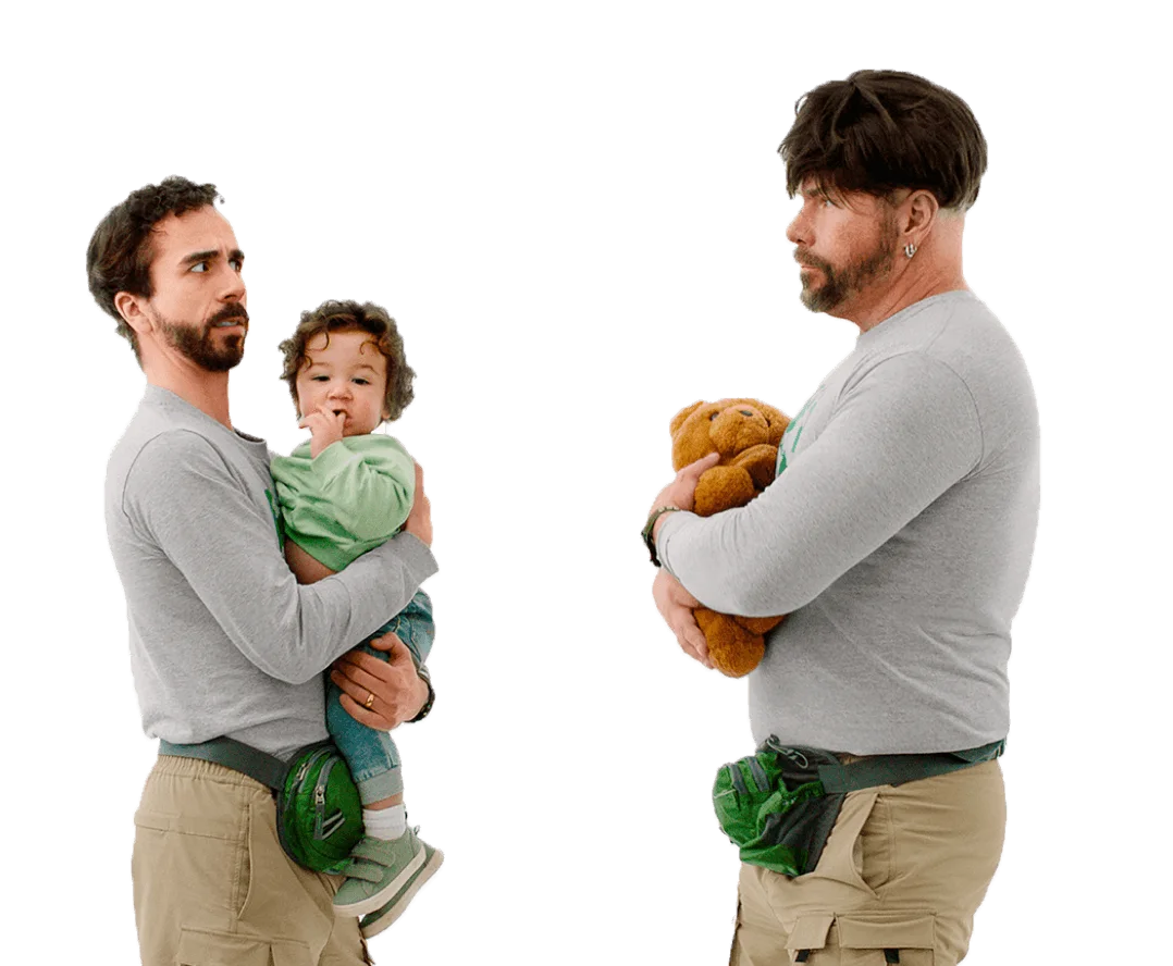 A man, holding a baby, looks suspiciously at an unconvincing impostor: a man who’s dressed just like him, wearing a wig and holding a teddy bear. 