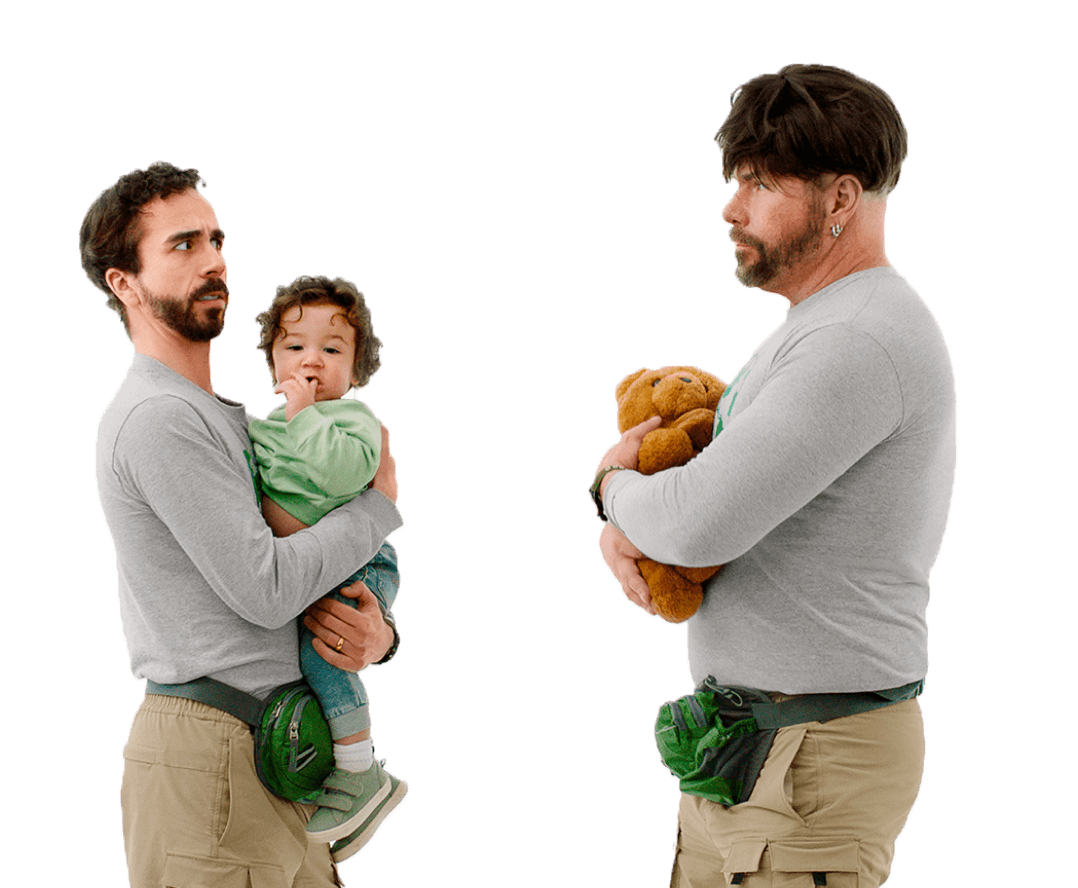 A man, holding a baby, looks suspiciously at an unconvincing impostor: a man who’s dressed just like him, wearing a wig and holding a teddy bear. 