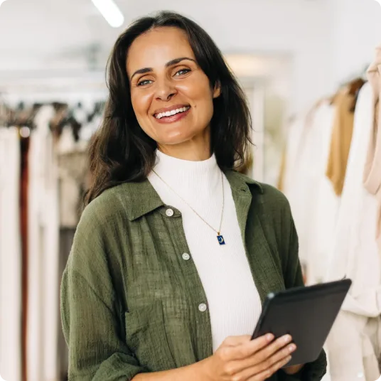 Retail owner smiles, holding a tablet among clothing racks.