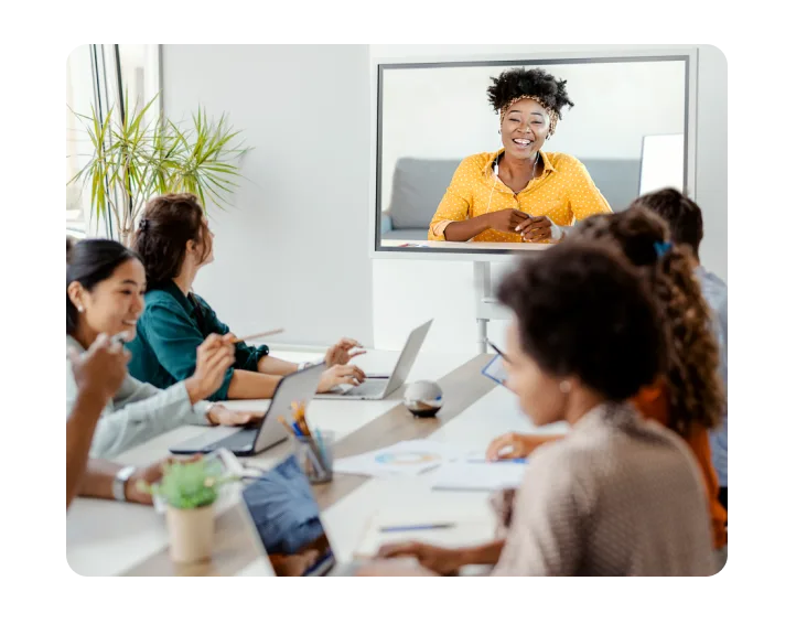 Group of six people in a conference room with open laptops, woman wearing yellow shirt, seated on a grey couch, on the videoconference screen. 