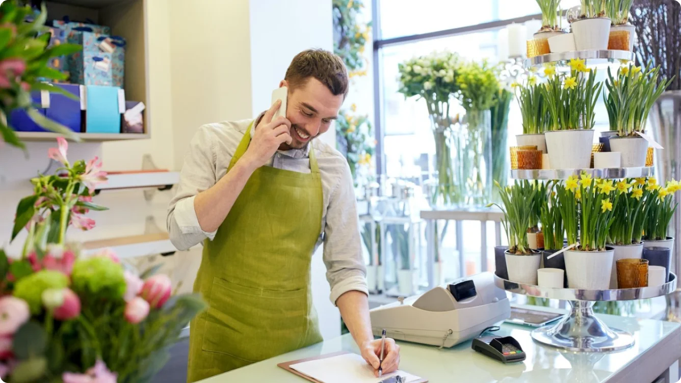 A smiling florist in a green apron talks on the phone and writes on a clipboard. The shop is filled with potted plants and flowers, with a cash register and card reader on the counter.