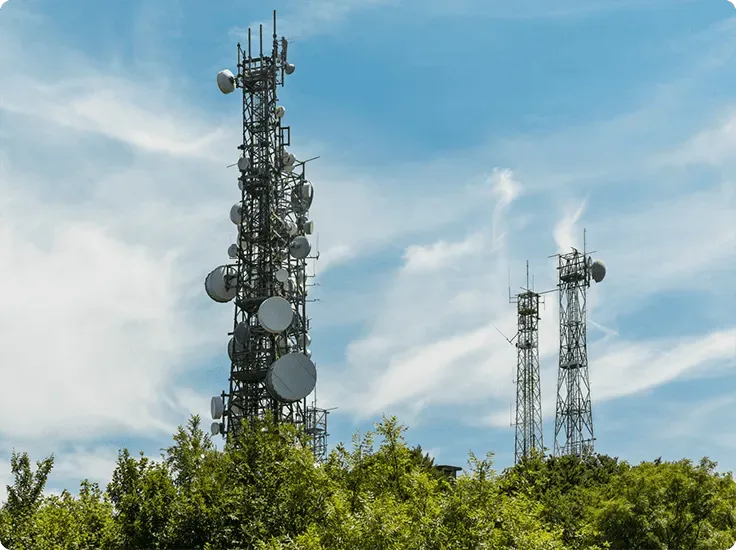 Cellular towers against a blue sky with light clouds and trees below.