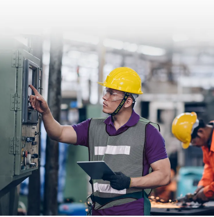 A person wearing a hard hat and vest working in a warehouse and using a tablet.