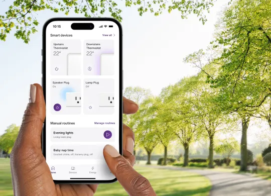 Une main tient un téléphone intelligent affichant l’interface de l’application Énergie intelligente, montrant des appareils intelligents et des routines manuelles sur un fond de parc ensoleillé.