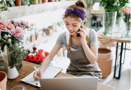 Un fleuriste souriant en tablier gris parle au téléphone et écrit sur une planchette à pince. La boutique est remplie de plantes et de fleurs en pot.