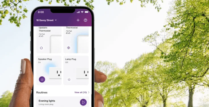 A hand holds the TELUS SmartHome+ app while out on a walk in the park showing the control of thermostats, smartplugs and routines.