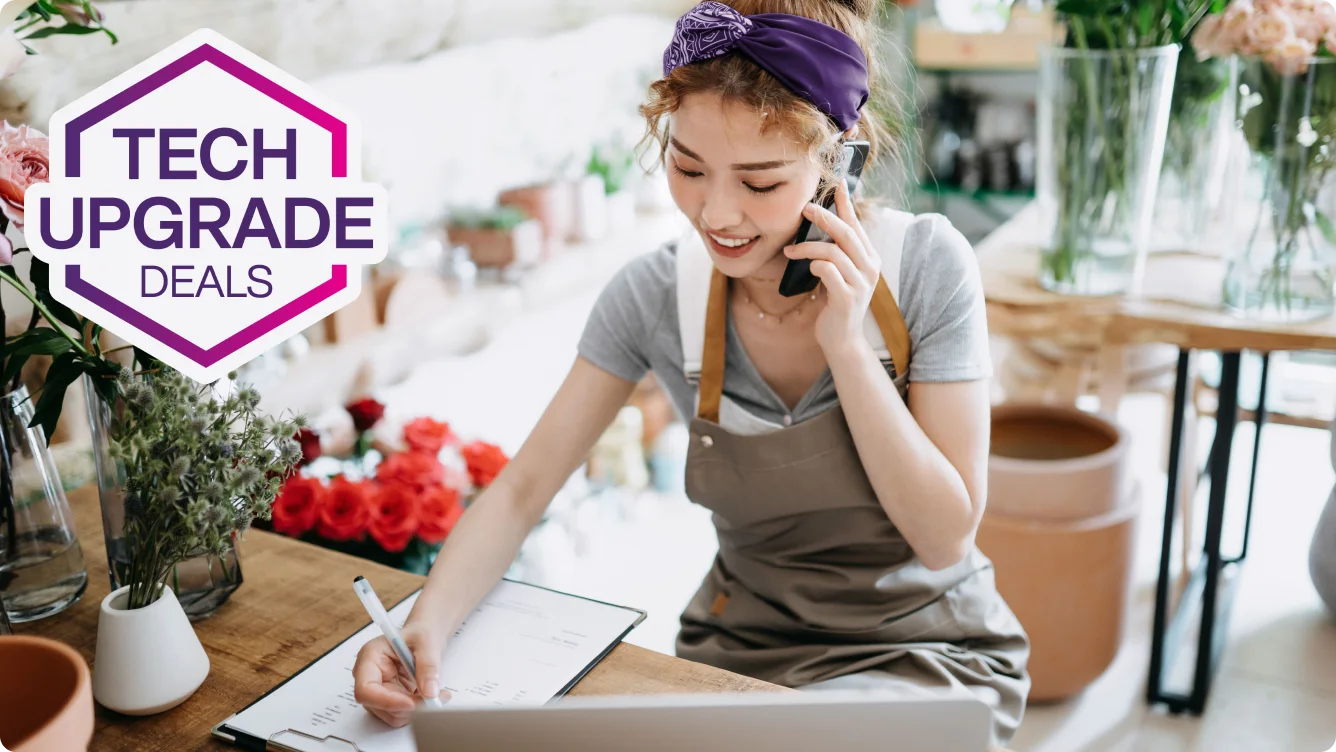 A smiling florist in a gray apron talks on the phone and writes on a clipboard. The shop is filled with potted plants and flowers. A roundel with the words “Tech Upgrade Deals”.