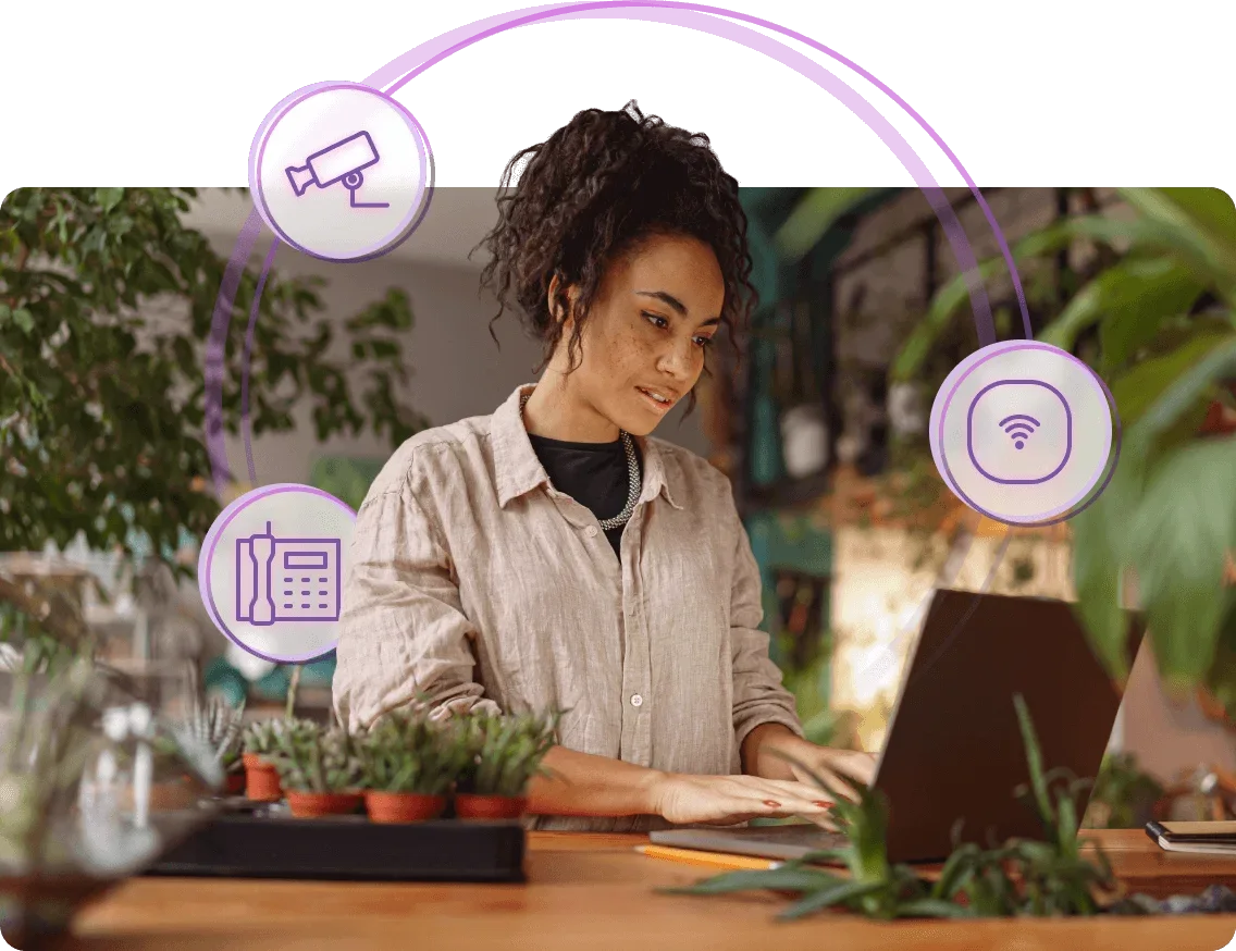 A business owner in a flower shop working at a laptop with office phone, camera and wi-fi icons around them.