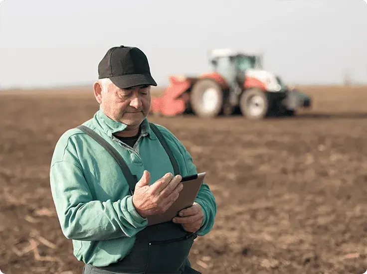 A man working in a field uses high-speed internet on a tablet with a tractor in the background.