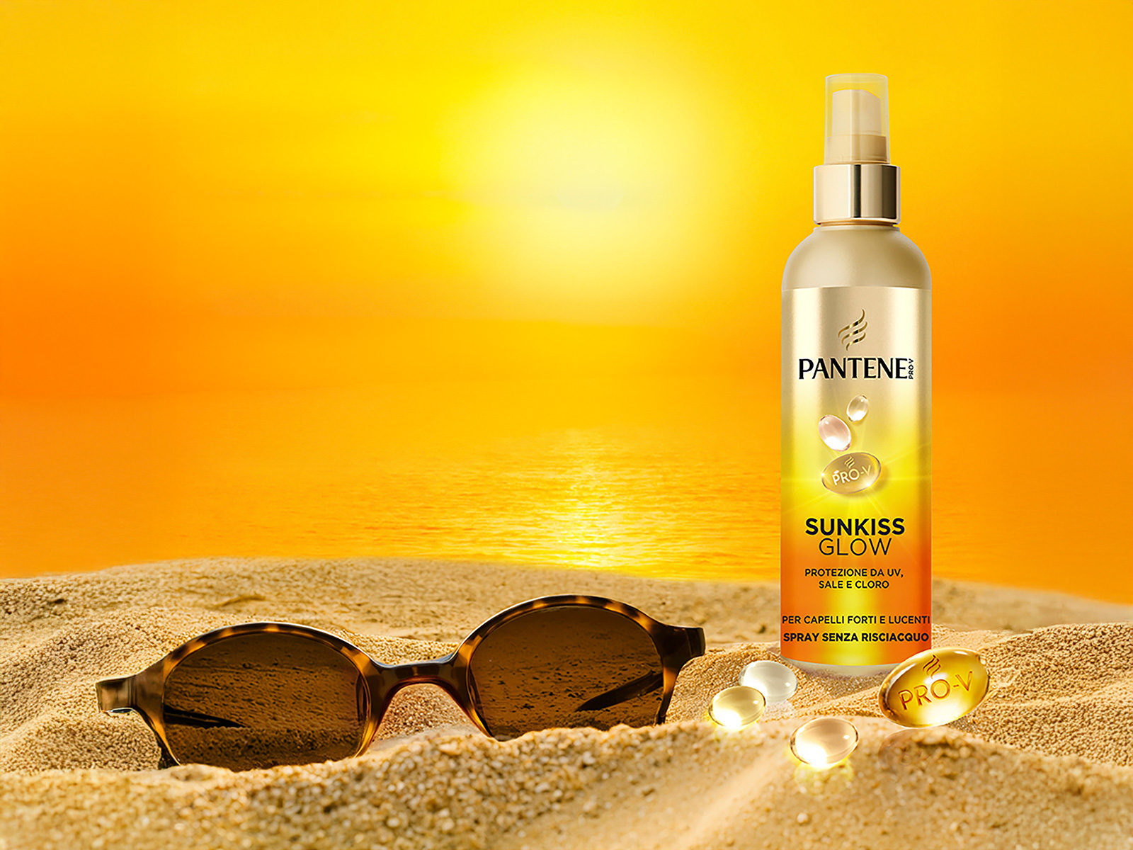 Pantene Spray Protezione Solare Sunkiss in piedi sulla spiaggia accanto agli occhiali da sole. Sullo sfondo un tramonto arancione.