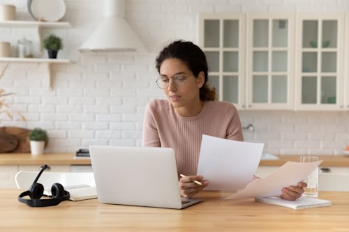 Thoughtful female sat at home workplace holding financial documents looking on computer screen.