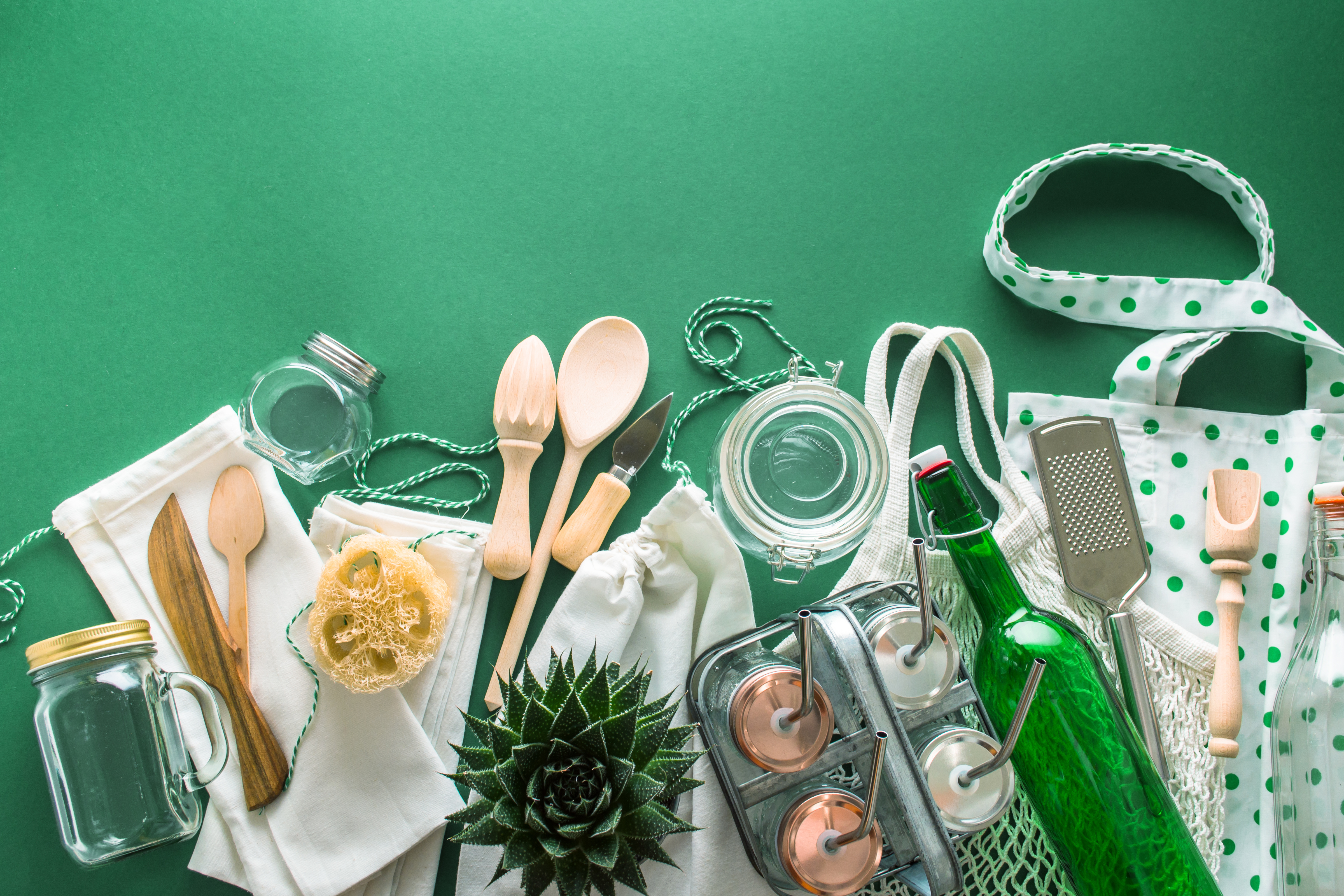 Grüner Hintergrund mit umweltfreundlichen Utensilien. Darunter Holzbesteck, Glasflaschen, Baumwoll-Tragetaschen und eine Pflanze.