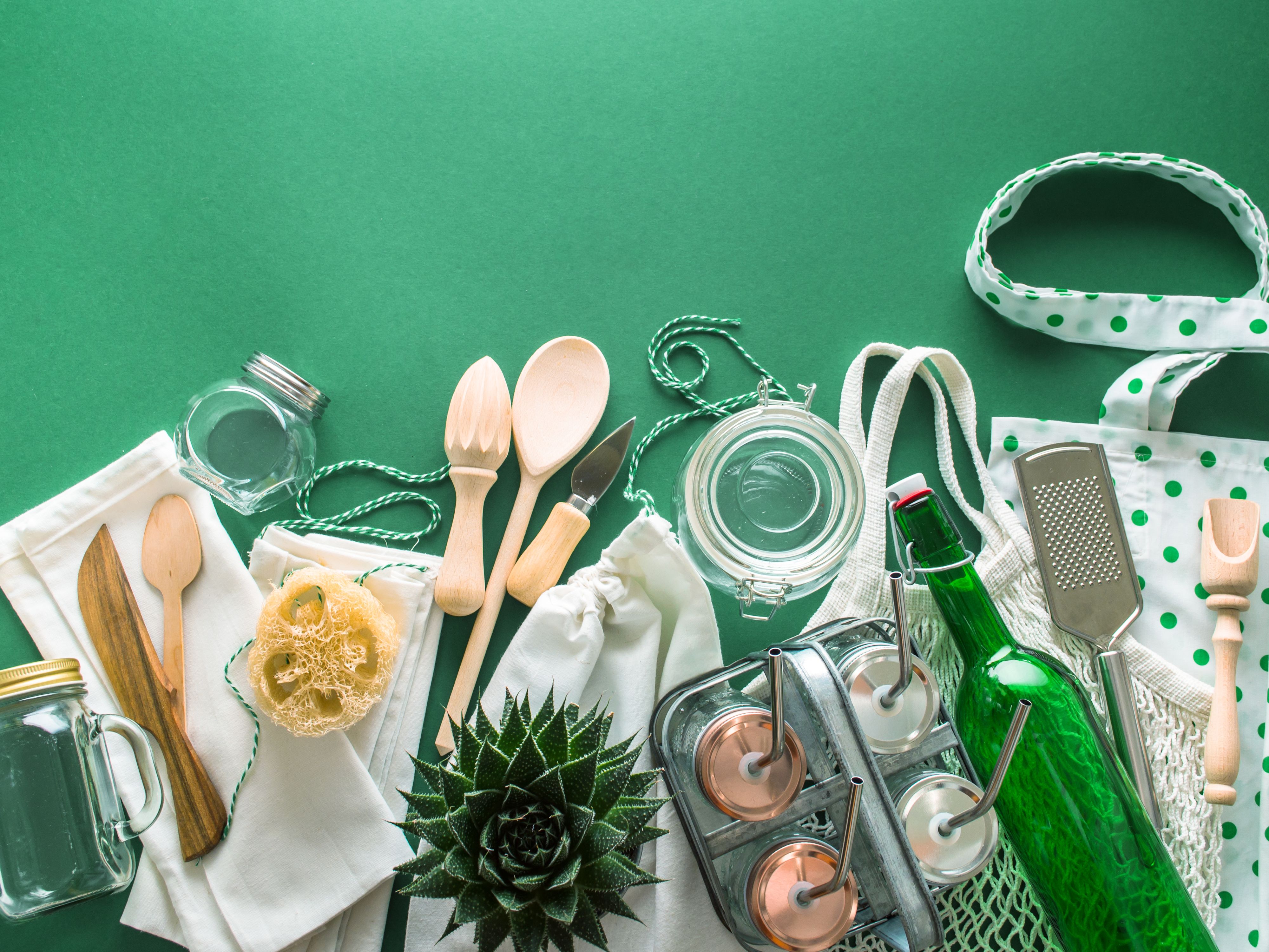 Grüner Hintergrund mit umweltfreundlichen Utensilien. Darunter Holzbesteck, Glasflaschen, Baumwoll-Tragetaschen und eine Pflanze.