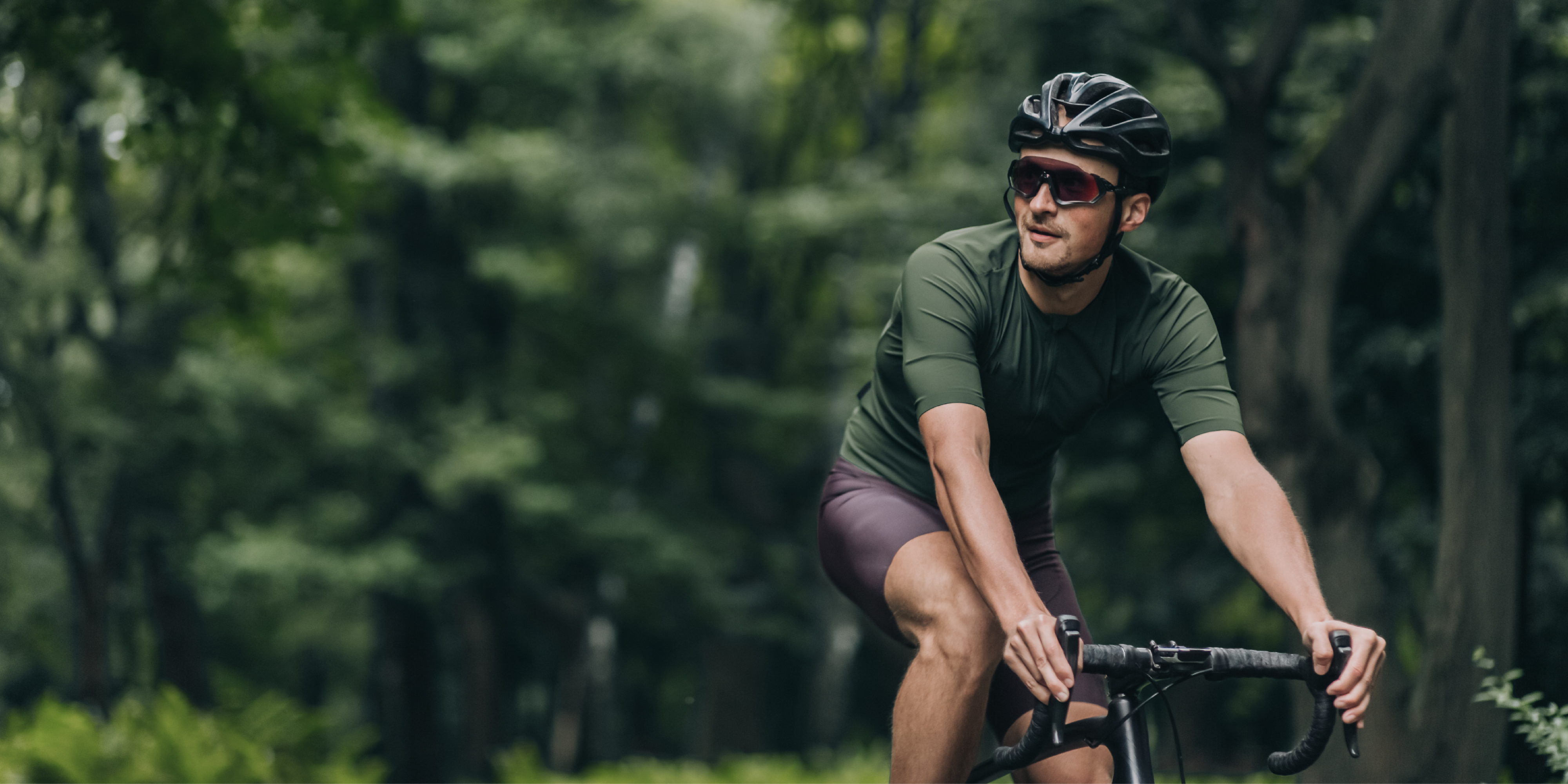 Ein in die Ferne lächelnder Mann, etwa Anfang dreißig, trägt einen Fahrradhelm sowie ein Sport-Shirt und Radlerhose, während er durch den Wald mit seinem Fahrrad fährt.