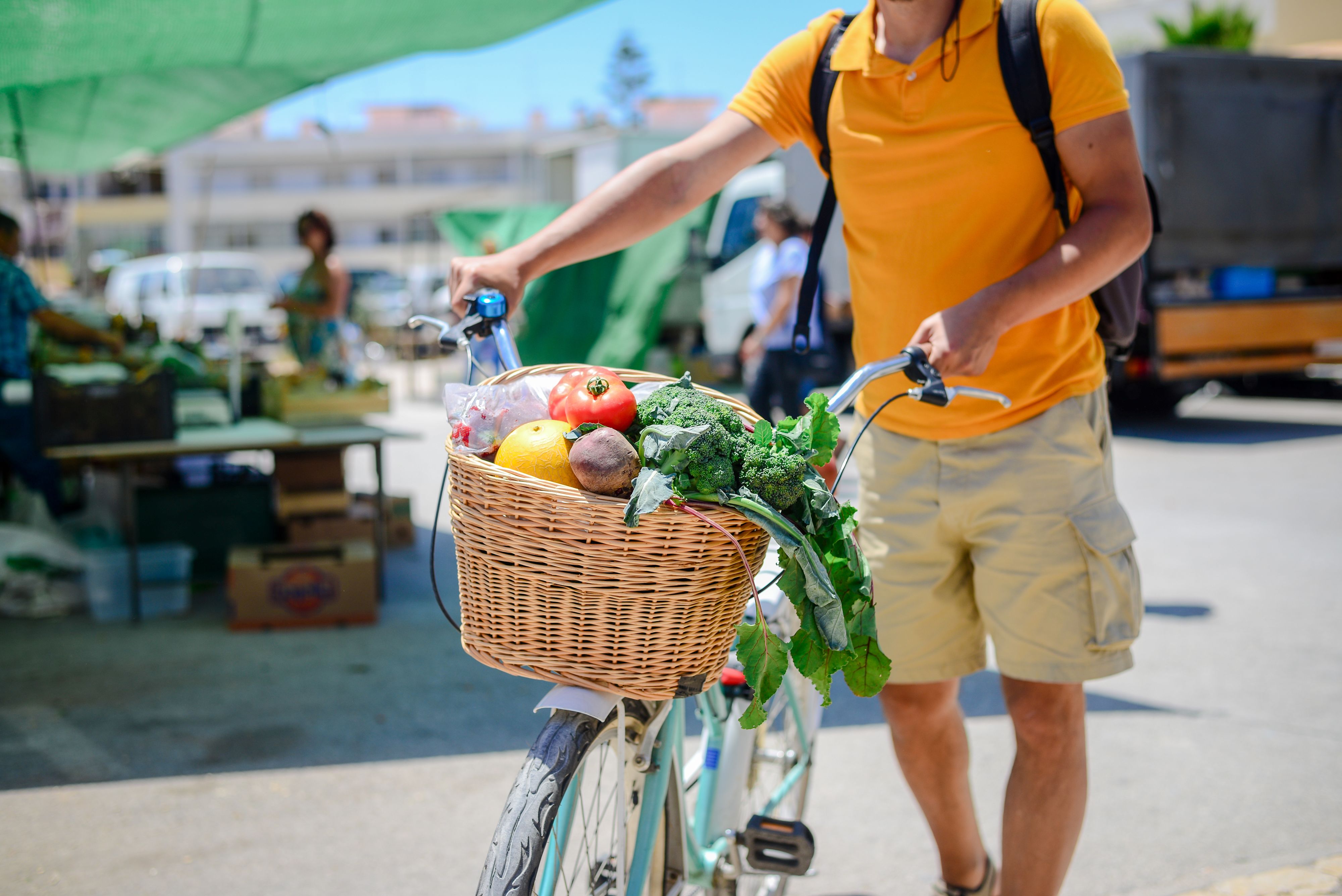 Mann der an einem sommerlichen Tag mit seinem Fahrrad über den Markt läuft und Obst und Gemüse im Fahrradkorb transportiert.