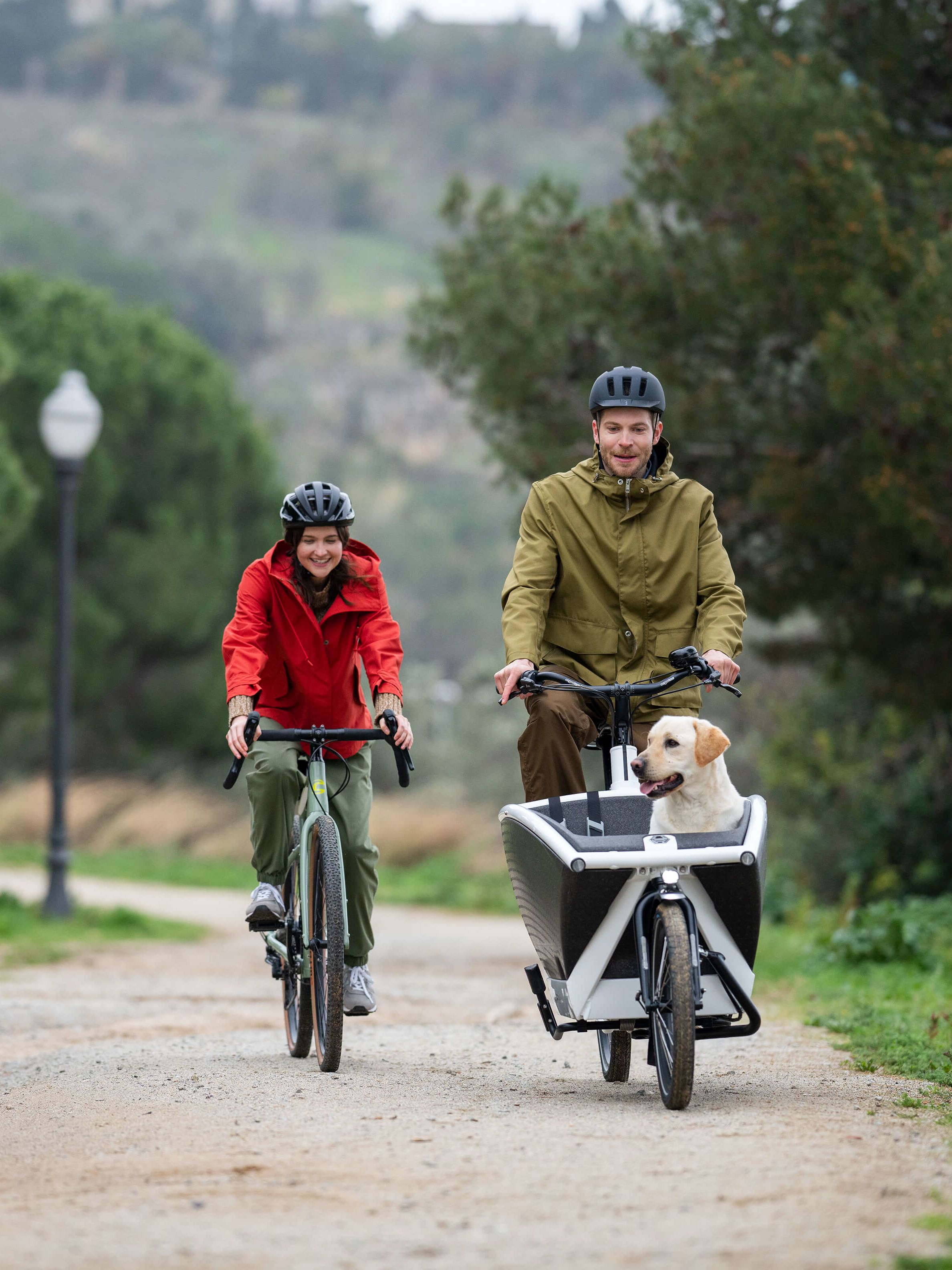 Zwei Menschen auf Fahrrädern. Aus dem Gepäckträger des einen Rades schaut ein Labrador heraus.
