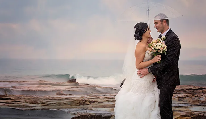 Bride and groom standing on the rocks by the ocean under an umbrella