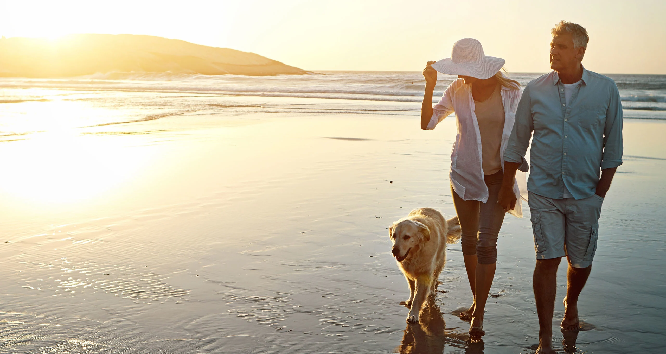 h man and woman walking along beach with golden at sunset
